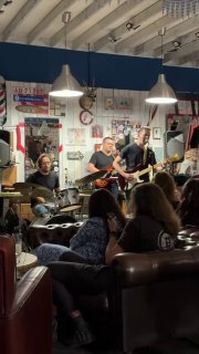 Джазовая группа на небольшой сцене внутри барбершопа Otto: музыканты с гитарой и ударными, публика сидит на диванах рядом со сценой.