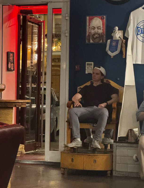 Мужчина сидит на импровизированном подиуме у входа в барбершоп Otto; вокруг видна тематическая атрибутика, портреты и мягкое освещение.