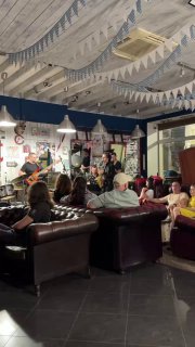 Широкий кадр зала барбершопа Otto во время джем-сессии: зрители на кожаных диванах, музыканты на сцене и гирлянды на потолке.