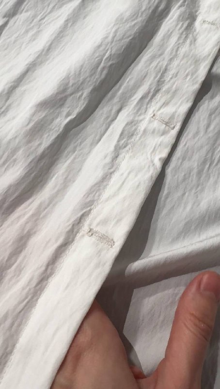 Крупный план белой ткани рубашки: видно строение ткани, швы и незавершённые петли — детали пошива и текстура материала под светом.