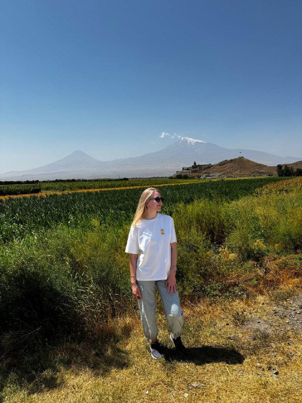 Автор стоящая на фоне горы Арарат: человек в светлой футболке на поле, позади виднеется заснеженная вершина и монастырский холм.