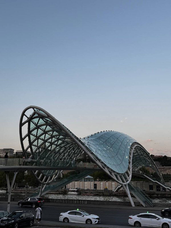 Панорамный вид на изогнутый стеклянно‑металлический пешеходный мост над рекой в Тбилиси в мягком вечернем освещении с движением машин внизу.