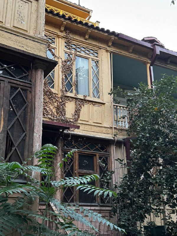 Деревянные резные балконы и фасады старого дома в Тбилиси: традиционная орнаментальная отделка, патина времени и густая зелень у фасада.