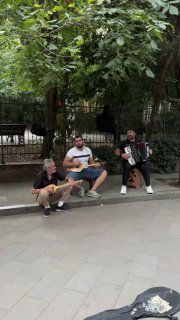 Уличные музыканты под деревьями в Тбилиси: аккордеон, гитара и барабан, прохожие наблюдают — сценка живой городской музыки и уличного перформанса.