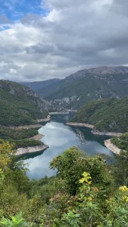 Панорамный вид на Пивское озеро: извилистая водная поверхность между высокими горными хребтами и растительностью на переднем плане под облачным небом.