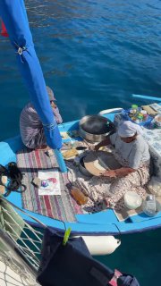 Небольшая лодка-продавец у борта яхты: двое людей готовят и раскладывают товары и еду на голубой лодке в спокойной бухте.