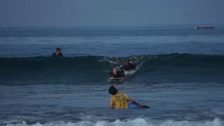 Сцена серфинга на спокойном пляже: серферы в воде ловят волну, видно несколько людей на досках и прибой — кадр про обучение и отдых на волнах.