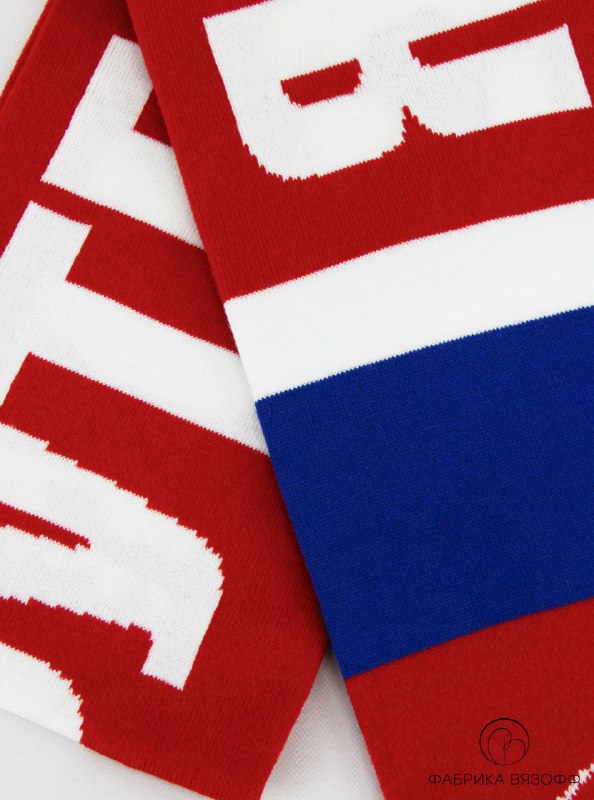 Крупный план вязаного полотна шарфа: фрагмент белых букв на красном фоне и полосы в цветах российского флага, видна текстура нитей и качество вязки.