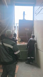 Работники в спецодежде направляют крупный блок плосковязальной машины через воротный проём в цех — этап установки и ввода оборудования в производство.