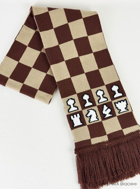 Шарф сложен и частично свернут; видна декоративная полоса с вязанными белыми и тёмными шахматными фигурами на фоне коричнево‑бежевой клетки.