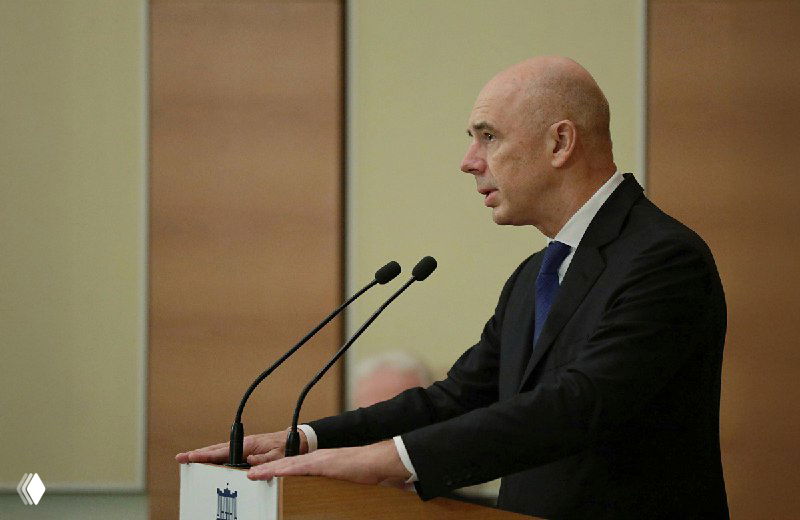 Мужчина в костюме выступает у трибуны на официальном совещании: два микрофона и нейтральный фон, кадр в профиль.