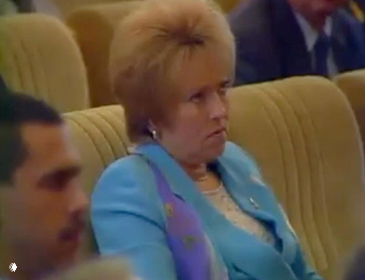 Архивное фото: женщина в светло-голубом пиджаке в зрительном зале, внимательное выражение лица, ряды слушателей на заднем плане.