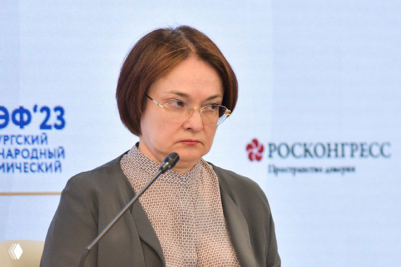 Женщина в деловом образе у микрофона на фоне баннера форума: серьёзное выступление, официальная обстановка и деловая аудитория за кадром