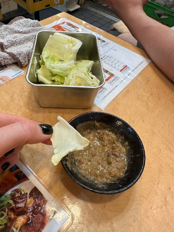 Белокочанная капуста в металлической коробочке рядом с миской кисло‑сладкого орехово‑соевого соуса: видно лист, готовый к маканию и рабочую поверхность стола.