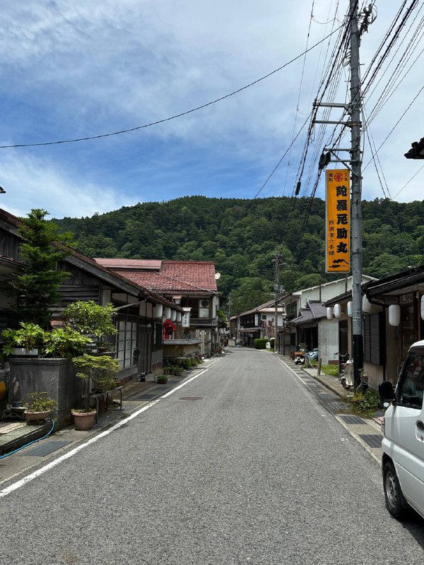 Пустая улица горной онсенной деревни Dorogawa Onsen: деревянные дома вдоль дороги и зелёные холмы на горизонте под голубым небом.