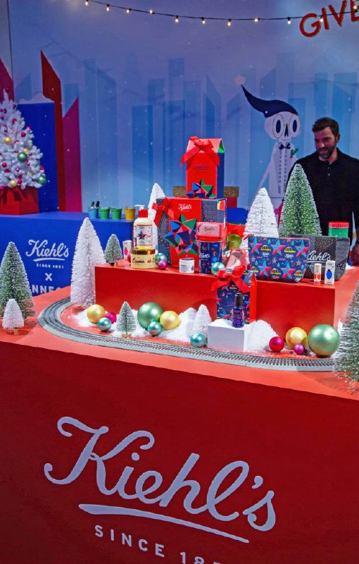 Праздничный экспозиционный стол Kiehl's с подарочной упаковкой, миниатюрными декорациями и атмосферной красной обивкой брендинга.