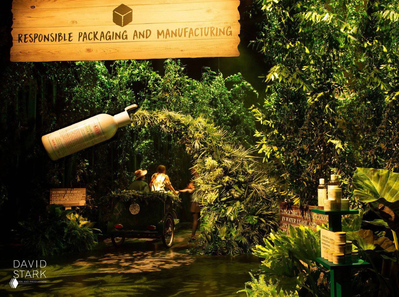 Тёмная тематическая инсталляция в стиле джунглей: густая зелень, вывеска Responsible packaging и большой парящий флакон косметики над сценой.