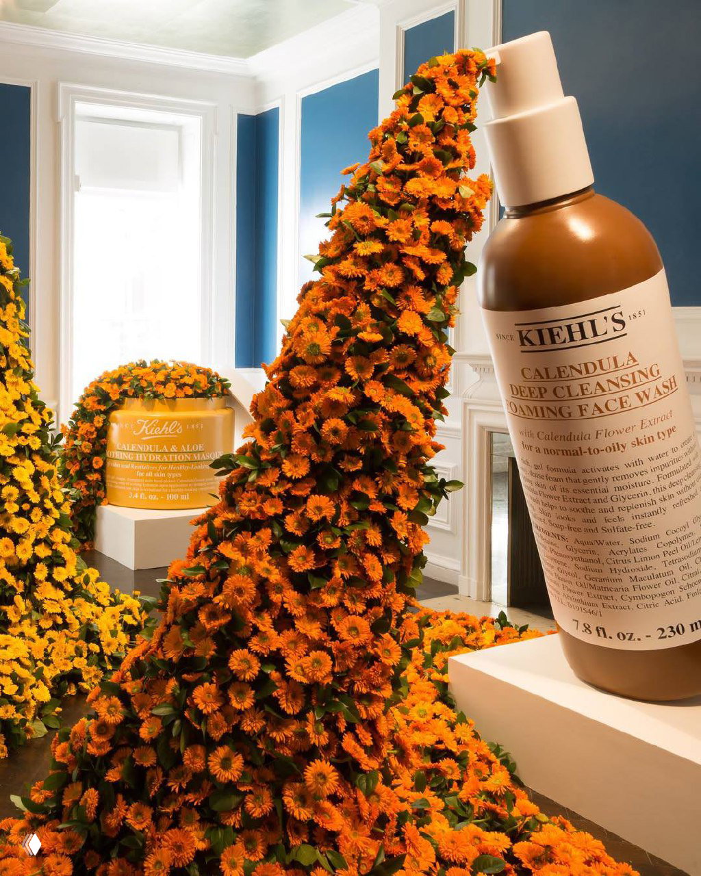 Брендовая инсталляция с большим флаконом Kiehl's и каскадом из оранжевой календулы: сочетание продукта и цветочной композиции в выставочном пространстве.