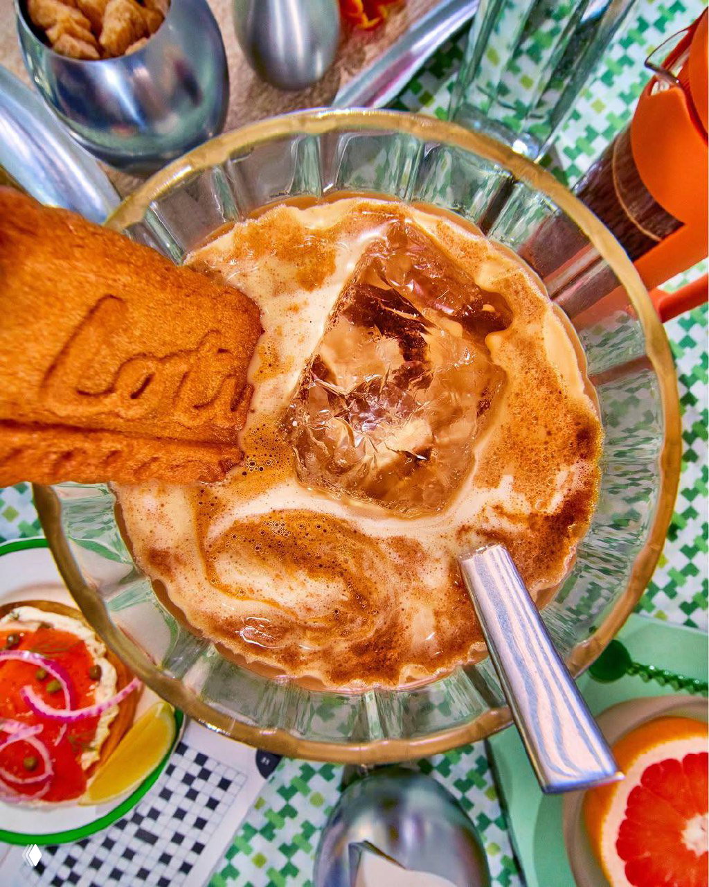 Вид сверху: стакан с кофейным напитком и большим кубиком «фейкового» льда, рядом кусочек печенья Biscoff, яркая сервировка на столе.