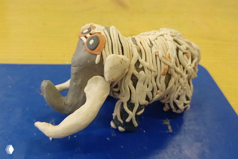 Мелкая скульптура мамонта из пластилина/скульптурного материала, текстурная поверхность и белые бивни, фотография детской поделки крупным планом.