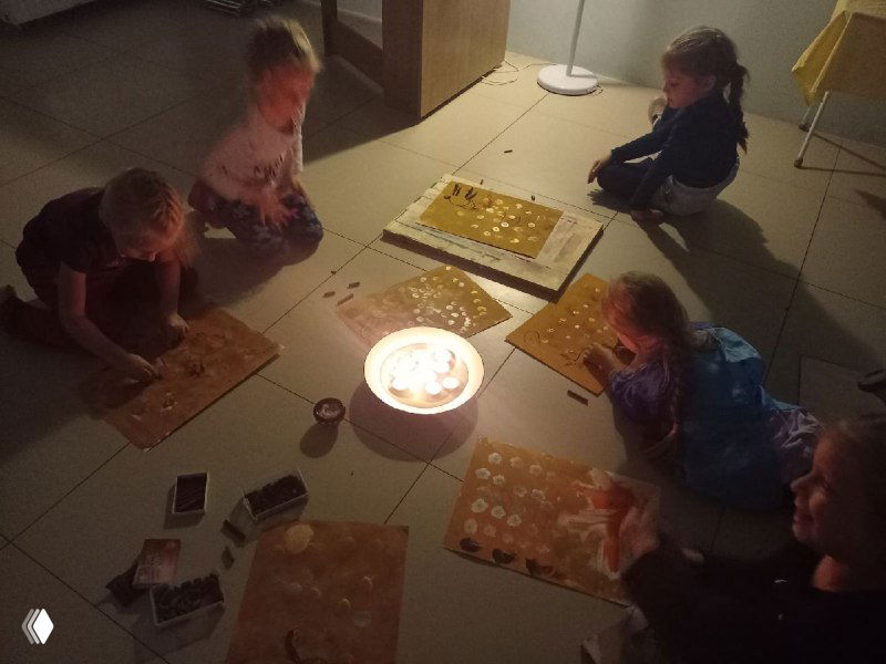 Вечернее занятие при свечах: дети рисуют на полу при тёплом свете свечи, атмосферная сцена с рабочими материалами и руками детей, фотография процесса.