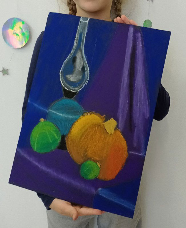 Ученица держит лист с натюрмортом сухой пастелью: вытянутая стеклянная форма лампы, яркие оранжевые и зелёные фрукты на ткани.