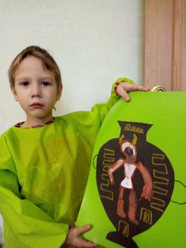 Ребёнок держит свою работу: изображение вазы с персонажем на зеленом картоне, видна гордая поза ученика и школьная обстановка.