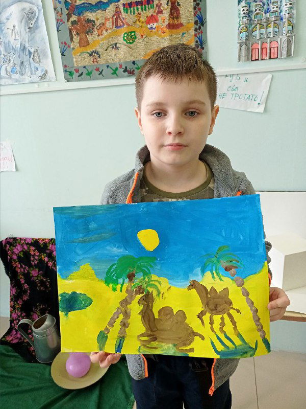 Мальчик демонстрирует картину с пальмами и сюжетной сценой каравана или сказочной истории, написанную яркими акриловыми или гуашевыми красками.