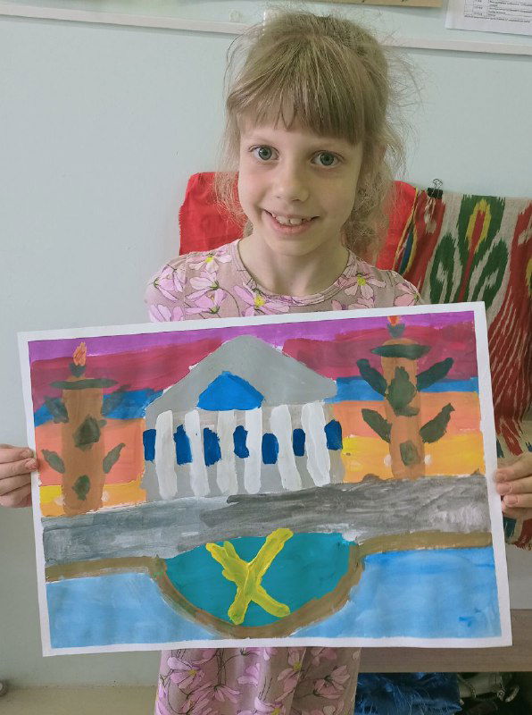 Ребёнок показывает плакатную работу с порталом и колоннадой; композиция выполнена яркими красками на фоне стен класса и декораций.