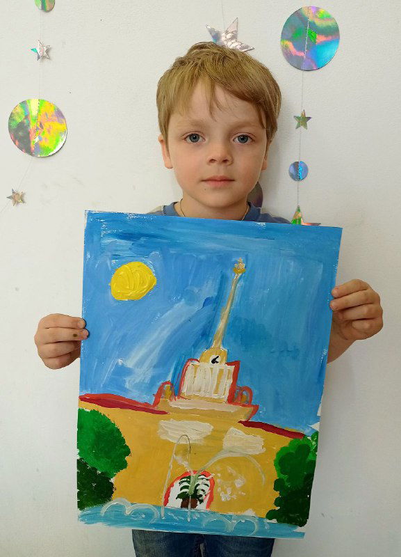 Мальчик в студии держит рисунок с вертикальным силуэтом здания и солнцем; видно простую детскую живопись и белую стену с декором.