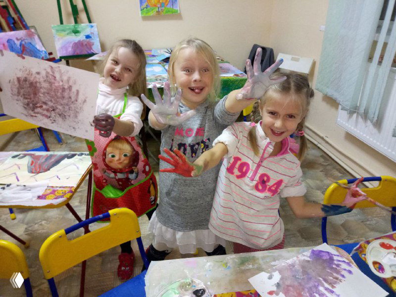 Трое девочек в классе рисования с грязными руками и столами, много бумажных листов с отпечатками ладоней и смешанными цветами вокруг.