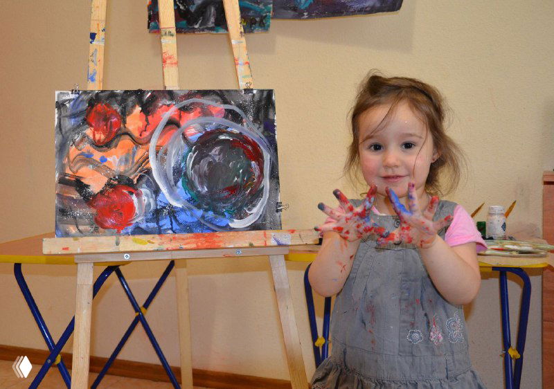 Маленькая девочка показывает ладони, испачканные краской, на мольберте рядом незаконченная картина — сцена творческой работы в классе.