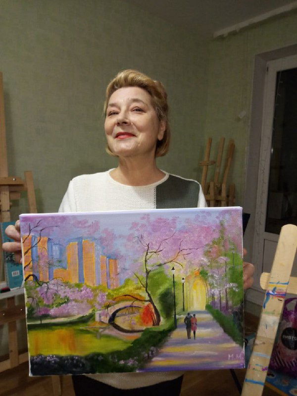 Пожилая участница улыбается и держит большой холст с городским парком и дорожкой, пастельные тона и декоративная подача в технике мастеркласса
