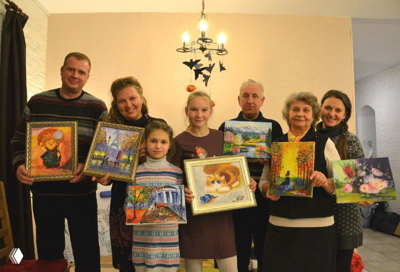 Несколько семей и участников разных поколений в группе демонстрируют оформленные картины и рамки — итоговый снимок группового мастер-класса в студии