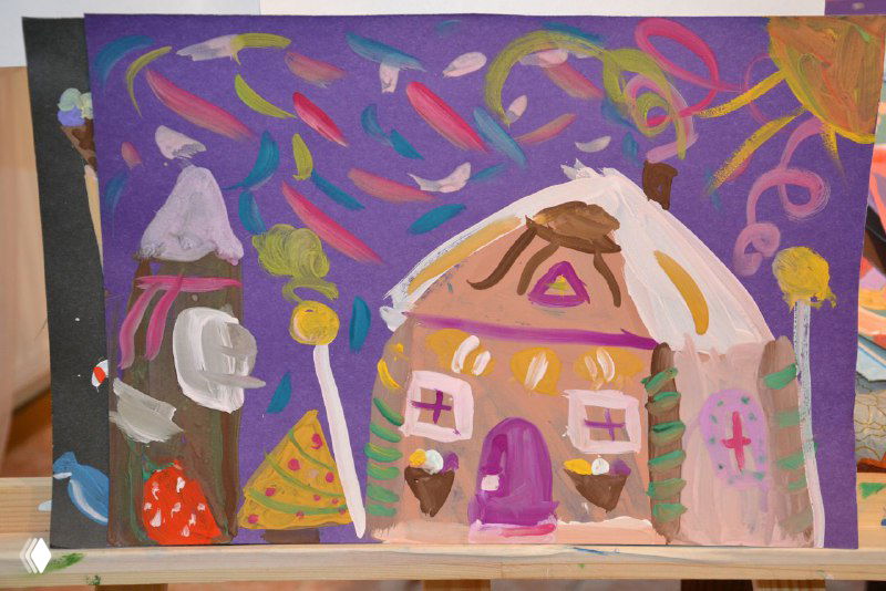 Работа ребёнка на мольберте: сказочный домик на фиолетово-розовом фоне с декоративными элементами и наивной перспективой.