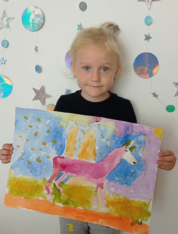Ребёнок держит картину с розовой лошадью на фоне декоративной стены студии, демонстрация результата занятия по рисованию для дошкольников.