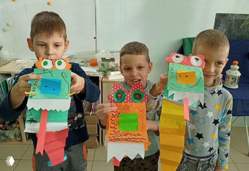 Трое мальчиков демонстрируют разноцветных бумажных драконов с большими "очками"-глазами, зубыми рядами и бумажными хвостами в мастерской.