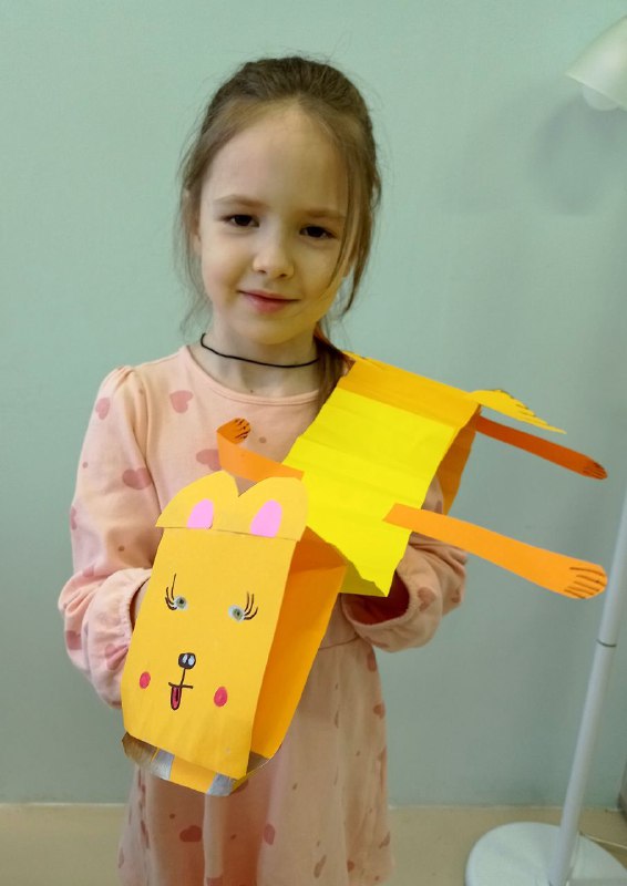Девочка держит длинного жёлто-оранжевого бумажного дракона с головой, сделанной буквой П, видно лапы и хвост из полосок бумаги.