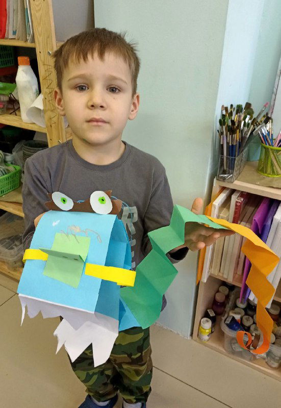 Мальчик с синим бумажным драконом: крупный план головы с приклеенными глазами, зубами и декоративными элементами, классная атмосфера.