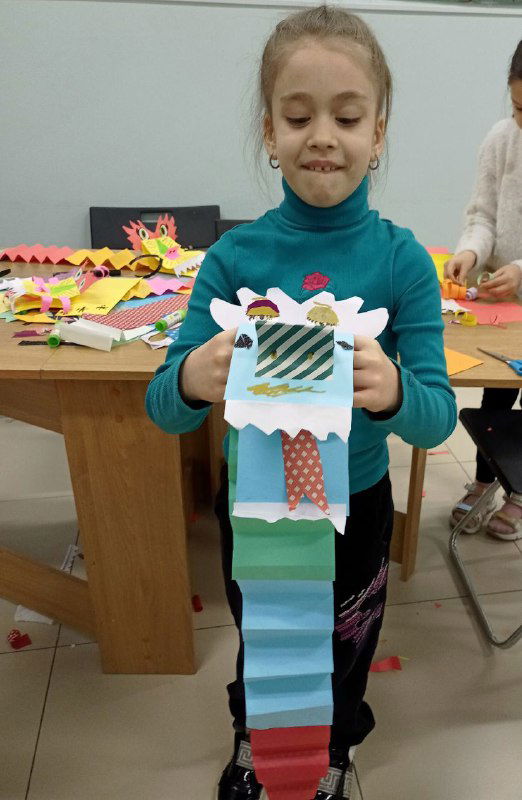 Девочка показывает бумажного дракона с гармошкой-туловищем и украшениями из полосок бумаги; рабочее место с материалами виднеется на фоне.
