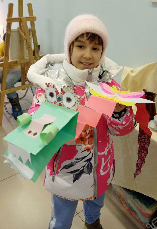 Ученик в шапке держит два бумажных дракона ярких оттенков: видны глазки, носы и декоративные элементы, снято в студии рисования.