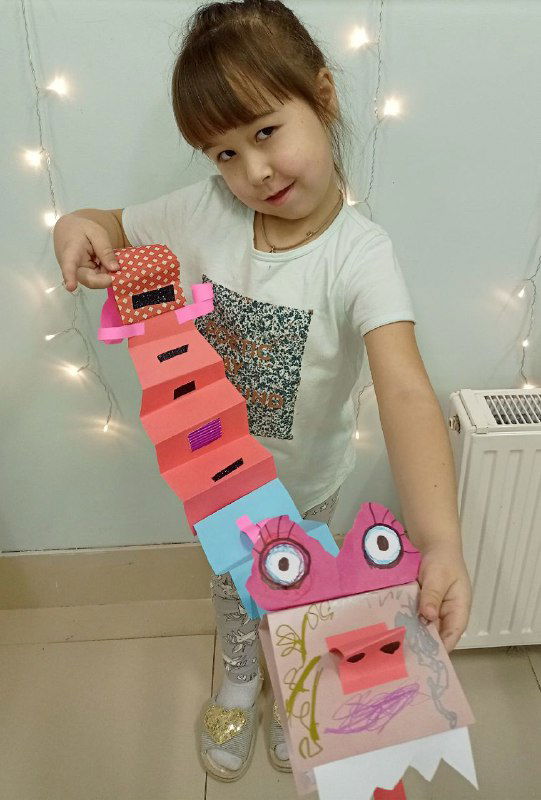 Девочка с розово-голубым бумажным драконом, видно детали головы, приклеенные глаза и зубы, а также декоративные элементы на туловище.