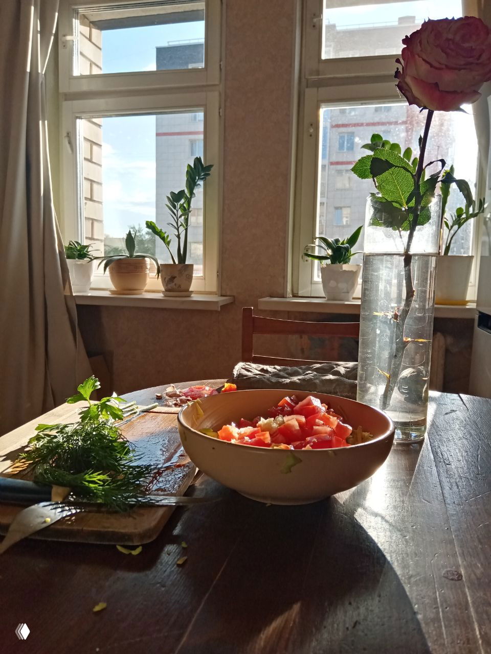 Уютный кухонный стол у окна с миской салата, рубленой зеленью на доске и розой в вазе — домашняя сцена подготовки еды при солнце.