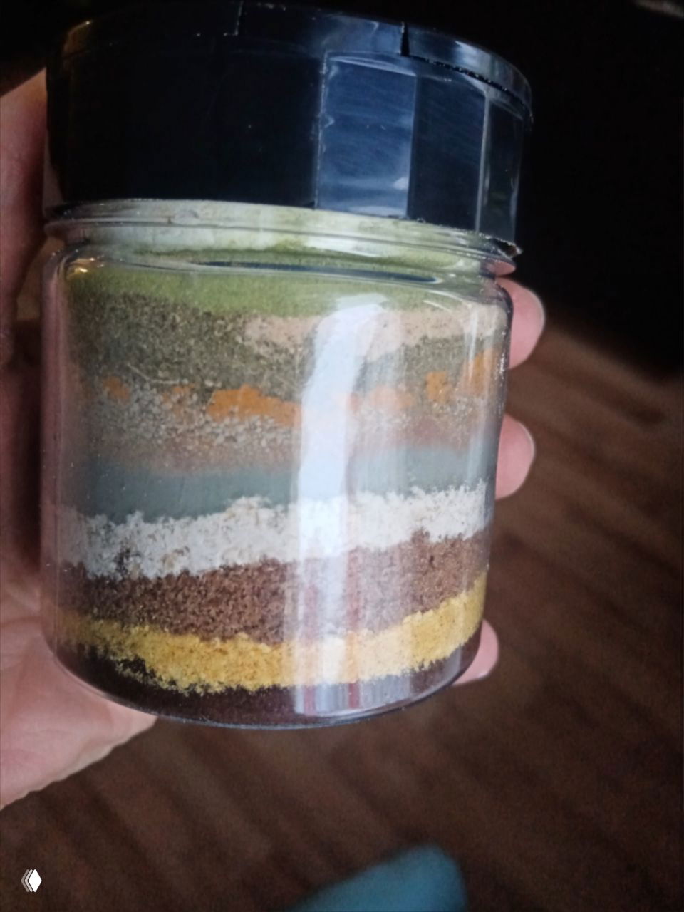 Крупный план стеклянной баночки с яркими разноцветными слоями убтана: зелёные, серые, белые, коричневые полосы; видна текстура и помол ингредиентов.