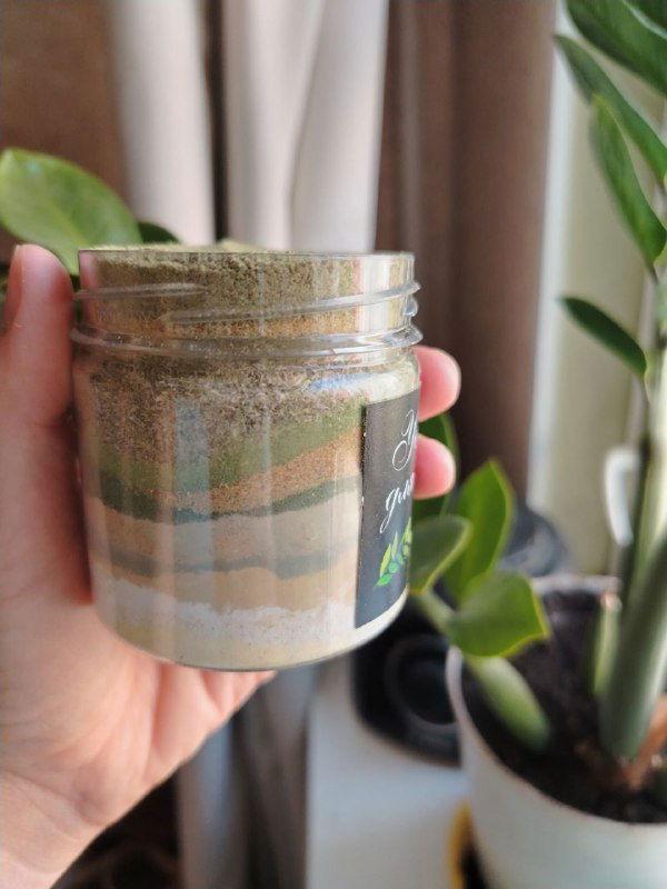 Стеклянная банка с многослойным порошковым убтаном в руке автора, видно текстуру и слои травных смесей на фоне домашнего интерьера.