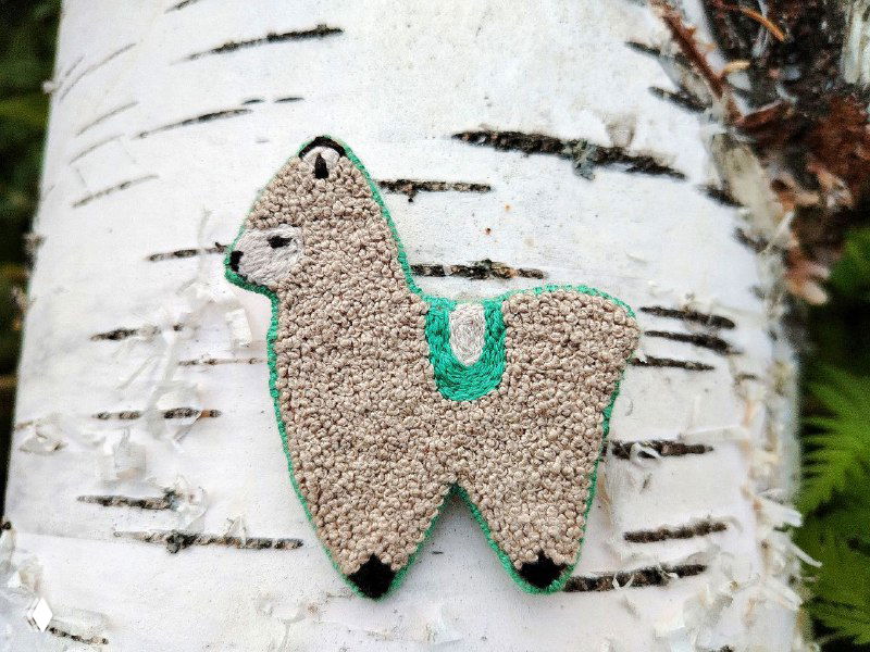 Другая версия броши‑ламы с зелёной окантовкой на фоне берёзовой коры; видно мелкие стежки, ровную отделку и контрастные детали.