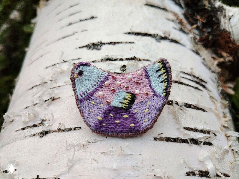 Пастельная брошь‑курочка фиолетовых и мягких тонов на коре берёзы; видны мелкие вышитые детали и аккуратные цветовые акценты.