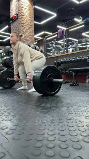 Женщина в тренажёрном зале выполняет становую тягу со штангой на резиновом покрытии; видны зеркала и тренажёры в фоне.