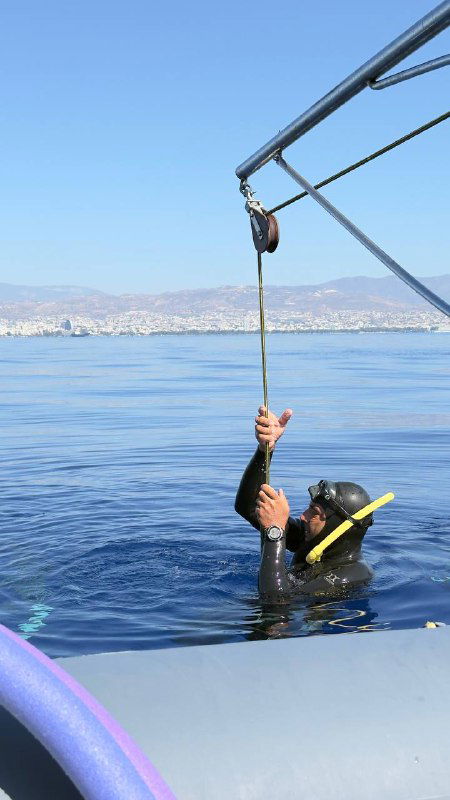 Ныряльщик на поверхности держится за трос рядом с лодкой, подготовка к глубокому или тренировочному погружению на фоне спокойного моря и побережья.