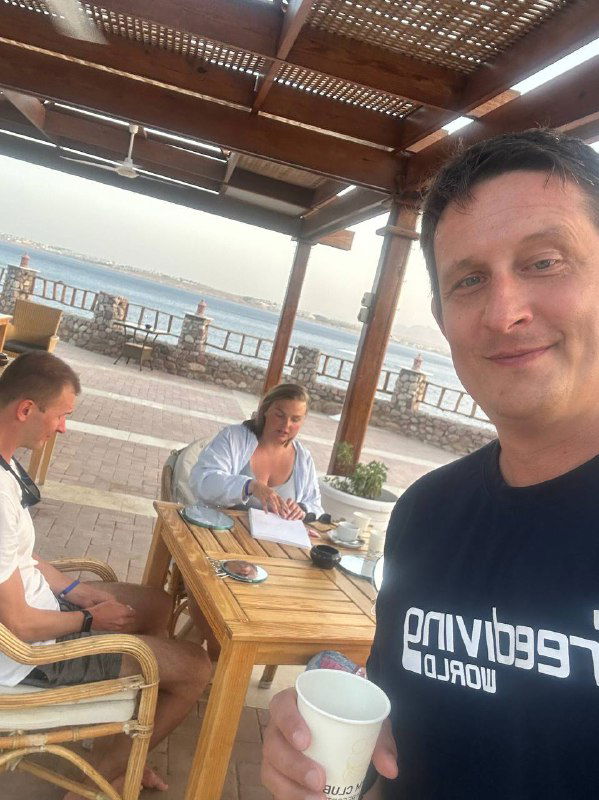 Селфи тренера на фоне учебной группы за столом: участники практикуют продувку, на заднем плане видна морская линия и прибрежная зона при заходе солнца